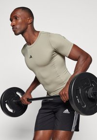 Těsné světle olivové sportovní tričko s krátkými rukávy, černé logo Adidas; černé šortky se třemi bílými pruhy na každé straně, držící činku.