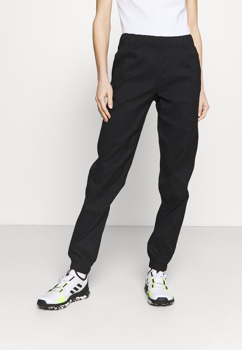 Personne portant un pantalon de jogging noir, des baskets blanches avec des accents noirs et jaune néon, et un t-shirt blanc, debout sur un sol blanc.