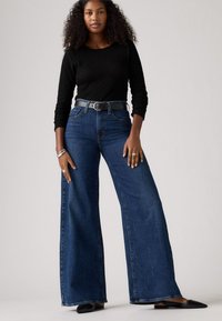 Jean en denim bleu taille haute, à jambes larges, avec une ceinture noire. Ajustés à la taille, ils ont une texture lisse et une coupe décontractée.