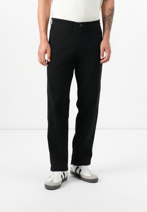 Model wearing black trousers and white t-shirt, standing on a plain background. Shoes are white with black stripes.