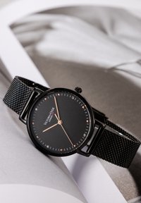 Czarny zegarek na czarnym siatkowym pasku z okrągłą czarną tarczą, różowo-złotymi akcentami, kryształkowymi znacznikami i teksturowaną powierzchnią. Nazwa marki widoczna na środku.