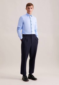 Camicia azzurro chiaro con bottoni in tessuto strutturato, abbinata a pantaloni plissettati blu scuro e scarpe nere. Sfondo neutro.