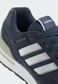 Adidas sporta apavi ar tumši zilas zamšādas augšdaļu, acs akcentiem, trim baltām svītrām un teksturētu gumijas zoli. Uz sāniem ir smalka zīmola identifikācija.