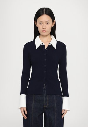 Marineblauer gerippter Cardigan mit einem weißen Kragenhemd. Er verfügt über eine Knopfleiste und lange Ärmel mit kontrastierenden weißen Bündchen.