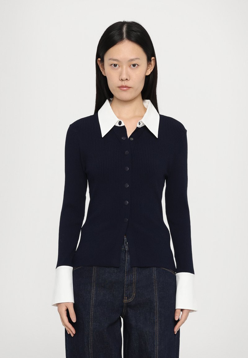 Donkerblauwe geribbelde cardigan met een witte, kraagloze blouse. Heeft een knoopsluiting aan de voorkant en lange mouwen met contrasterende witte manchetten.