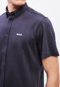 Tumesinine lühikeste varrukatega polo särk, valmistatud siledast kangast, millel on nööbitav krae. Rinnaesisele on trükitud diskreetne valge "BOSS" logo ning tumedad nupud.