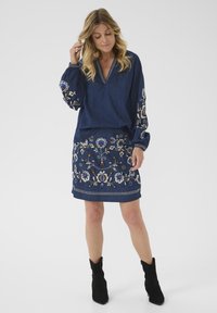 Haut en denim avec de longues manches bouffantes, ornée d'une broderie florale complexe en bleu et crème. Accompagné d'une jupe en denim assortie brodée.