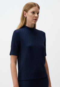 Marineblauw kortmouwtop met hoge hals, voorzien van een gestructureerd patroon. Gemaakt van een soepele stof, heeft het een relaxte pasvorm en een subtiele glans.