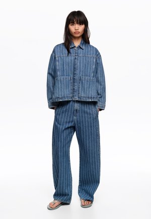 Kobieta stojąca, ubrana w niebieską, wąsko prążkowaną, oversize'ową kurtkę dżinsową i dopasowane szerokie spodnie z dżinsu, na szarych klapkach, na białym tle.