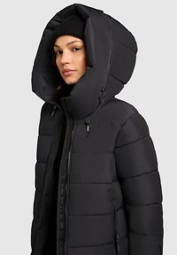 Čierna prešívaná bunda s veľkou kapucňou, ktorá má prešívané časti a predný zips. Textúrovaná tkanina, dve bočné vrecká a nastaviteľné šnúrky.