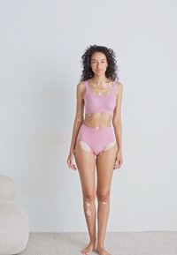 Femeie cu vitiligo, stând desculță, îmbrăcată în chiloți fucsia cu talie înaltă și un top scurt asortat, pe un fundal simplu.