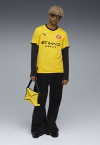 Gul sports t-shirt med sort kant, der har teksten og logoet "ETIHAD AIRWAYS". Bag det er der bredbenede sorte bukser og en gul taske.