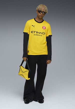 Gul sports t-shirt med sort kant, der har teksten og logoet "ETIHAD AIRWAYS". Bag det er der bredbenede sorte bukser og en gul taske.