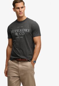 Graues Baumwoll-T-Shirt mit kurzen Ärmeln und aufgedrucktem "SUPERDRY & CO"-Logo. Kombiniert mit beiger Hose und einem braunen geflochtenen Gürtel, ergänzt durch eine Armbanduhr.