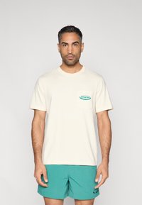 Μπεζ βαμβακερό t-shirt με κοντά μανίκια, διακοσμημένο με ένα πράσινο πέτσωμα με λογότυπο σε σχήμα κουτιού στο στήθος. Συνδυάζεται με τιρκουάζ μαγιό βερμούδα.