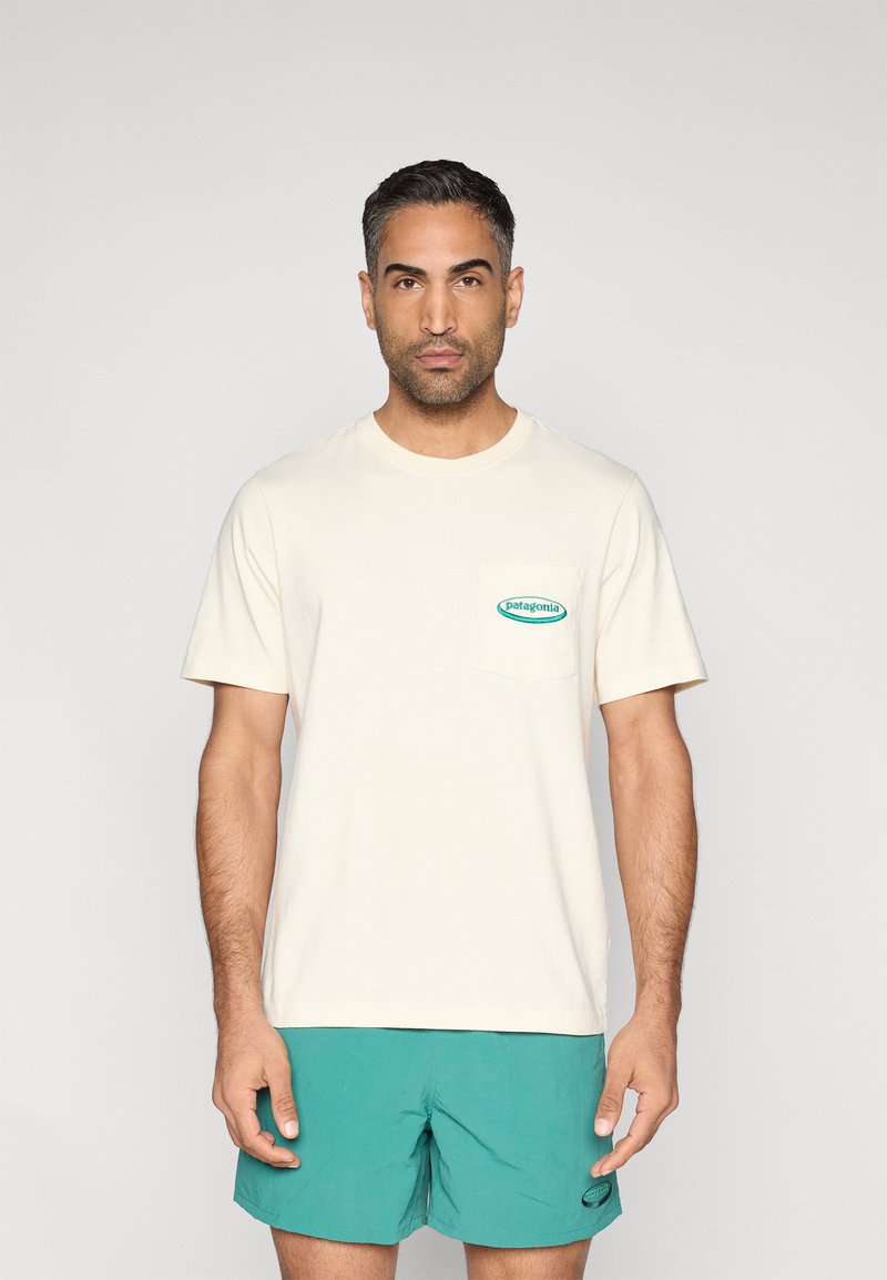 Μπεζ βαμβακερό t-shirt με κοντά μανίκια, διακοσμημένο με ένα πράσινο πέτσωμα με λογότυπο σε σχήμα κουτιού στο στήθος. Συνδυάζεται με τιρκουάζ μαγιό βερμούδα.