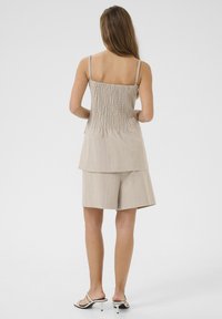 Top senza maniche beige chiaro con schiena arricciata, design a strati, abbinato a pantaloni corti coordinati. Il modello indossa sandali neri con cinturini.