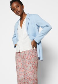 Blazer longo azul claro sobre uma blusa branca, combinado com calças plissadas estampadas que apresentam designs florais em vermelho e roxo.