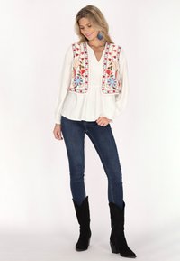 Witte blouse met bloemgeborduurde vest, gecombineerd met donkere spijkerbroek en zwarte cowboylaarzen. Heeft blauwe accenten en luchtige mouwen.