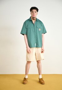 Grønn kortermet skjorte, beige shorts, brune sko. Skjorten har to frontlommer og et logopatch. Står på et sennepsfarget gulv.
