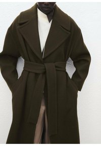 Manteau long vert foncé avec un large col, taille ceinturée et deux poches latérales, fabriqué en tissu texturé. En dessous, un pull léger est visible.