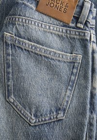 Svijetloplave traperice s lisnatom džepom na stražnjoj strani. Tkanina ima teksturirani, izblijedjeli uzorak. Na pojasu se nalazi kožni brend tag.