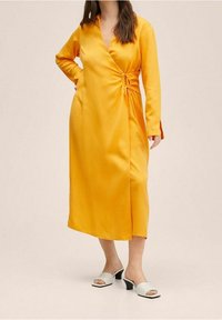 Gelbes Satinkleid zum Wickeln mit langen Ärmeln, seitlicher Raffung und knielangem Schnitt; kombiniert mit weißen offenen Mules.