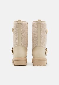 Beige ankle boots with a fuzzy shearling upper, smooth tan leather accents, decorative side buckles, and a textured rubber sole.