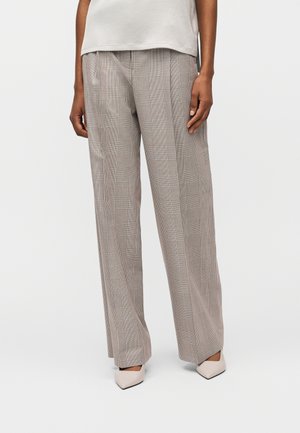 Osoba nosiaca vysoké široké kárované nohavice v béžových tónoch a špicaté béžové topánky, stojaca pred jednoduchým svetlým pozadím.