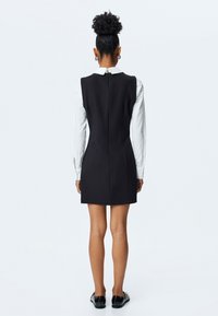 Schwarzes ärmelloses Kleid mit hohem Ausschnitt und Reißverschluss auf der Rückseite, getragen über einem weißen Langarmshirt. Glatte Textur mit taillierter Passform.