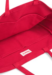 Röd canvas tote-väska med dubbla handtag, med en rymlig insida och en etikett sydd i botten. Inga synliga mönster eller dekorationer.
