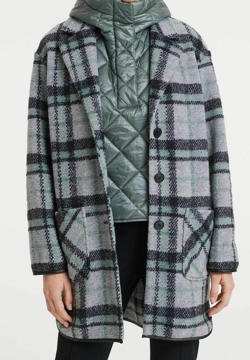 Personne portant un manteau en laine à carreaux gris et vert par-dessus une veste matelassée verte à col montant et un pantalon noir.