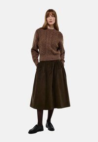 Pull en tricot marron avec un motif texturé, associé à une jupe plissée verte foncée, arrivant aux genoux. Des chaussures noires à lacets complètent le look.