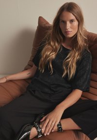 Black textured top with short sleeves, paired with loose black trousers and shiny black shoes featuring studded accents, sitting on a brown couch.