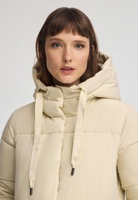 Beżowa kurtka puchowa z wysokim kołnierzem, kapturem i markowymi sznurkami ściągającymi. Charakteryzuje się pikowanym wzorem i elegancką sylwetką, zaprojektowana z myślą o ciepłu.