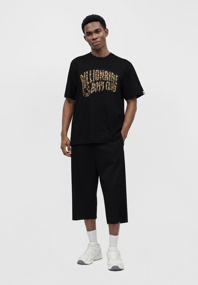 Νέος άνδρας που φοράει μαύρο γραφικό t-shirt, φαρδιά μαύρα κ cropped παντελόνια, λευκές κάλτσες και λευκά αθλητικά παπούτσια, στέκεται μπροστά σε μια απλή φόντο.