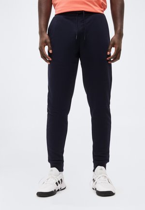 Man die marineblauwe joggerbroek, witte sportschoenen en een oranje shirt draagt, staand met ontspannen handen langs zijn zij tegen een effen achtergrond.