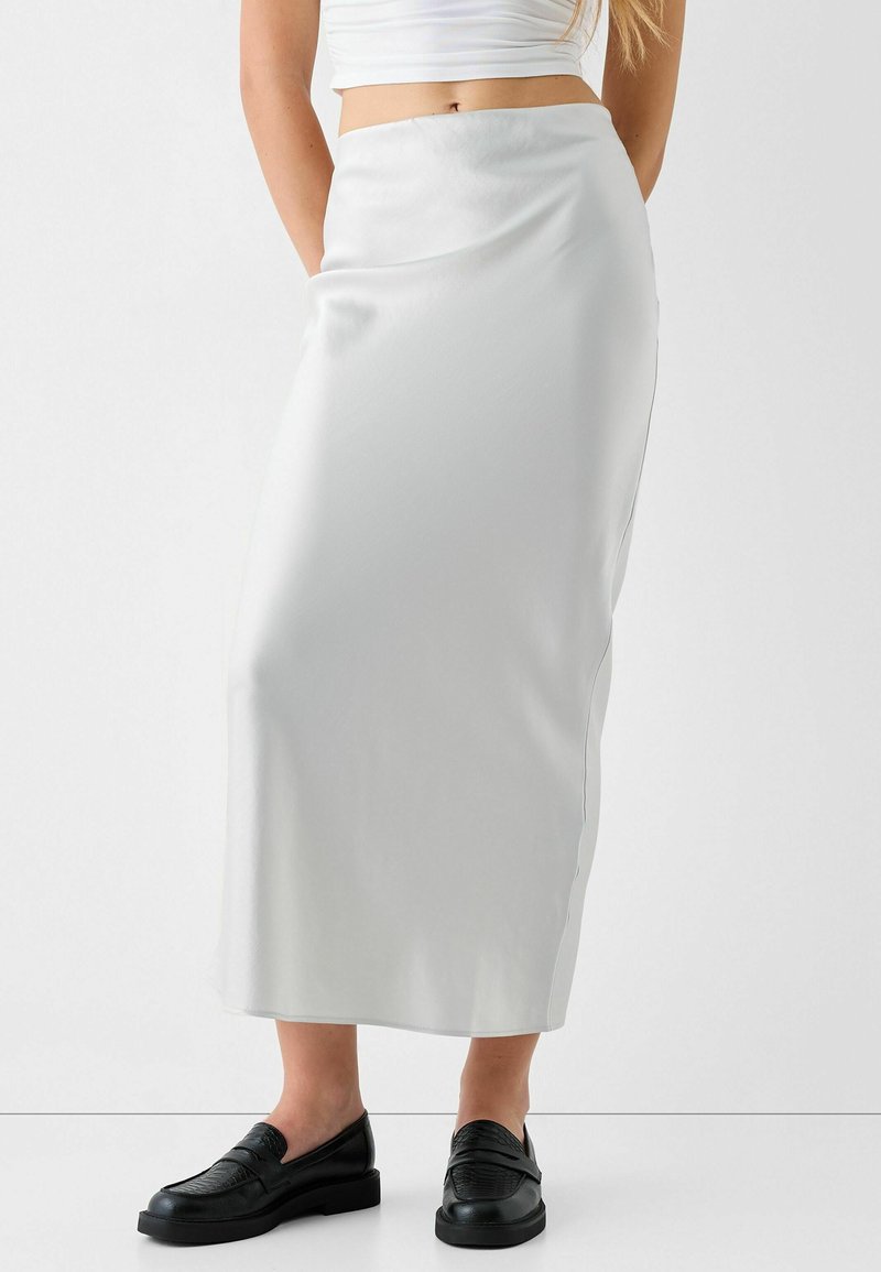 Bílá saténová maxi sukně s hladkou texturou, vypasovaným pasem a postranními kapsami, doplněná černými loafers s texturovaným vzorem.