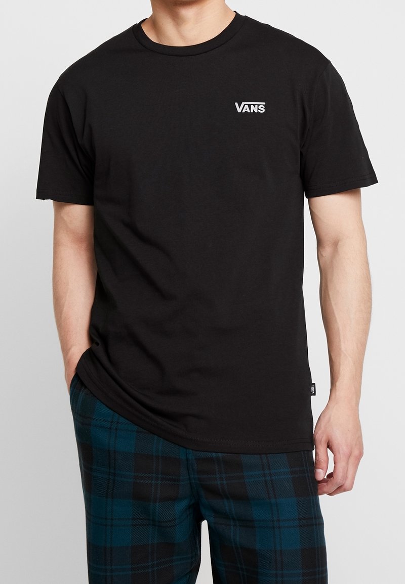Homme portant un t-shirt Vans noir avec un petit logo sur la poitrine et un pantalon à carreaux bleu-noir, les mains détendues le long du corps.