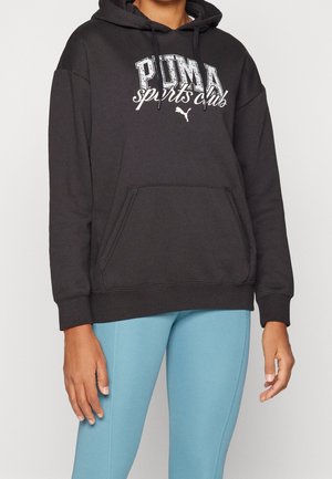 Osoba ubrana w czarną bluzę sportową Puma z kapturem i kieszenią kangurka oraz jasnoniebieskie legginsy, stojąca z rozluźnionymi ramionami.