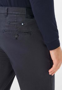 Pantaloni grigi scuri con una texture liscia; presentano due tasche posteriori con chiusura a pulsante e un'etichetta del marchio sulla cintura.