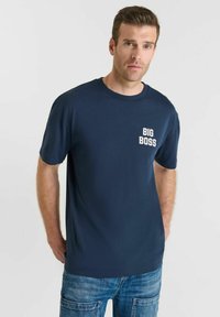 Man met kort bruin haar, gekleed in een marineblauw "BIG BOSS" t-shirt en blauwe cargobroek, staande met de handen achter zijn rug.