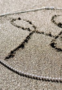 Perlenkette mit kleinen Perlen, die in einer gebogenen Linie auf einer sandigen Oberfläche angeordnet sind, umgeben von einem mit dem Finger gezeichneten Muster im Sand.