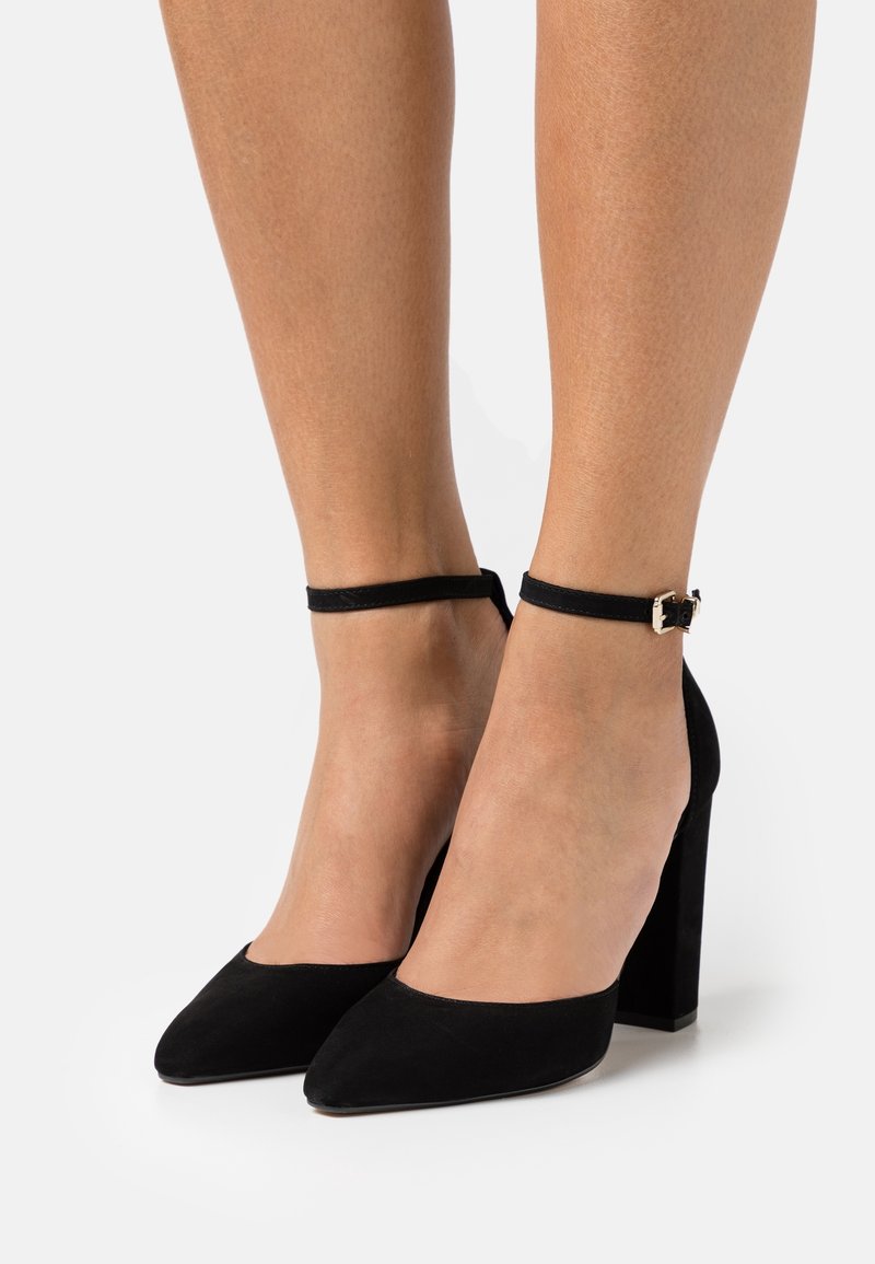 Chaussures à talons hauts en daim noir avec brides à la cheville et boucles dorées portées sur pieds nus sur un fond uni.