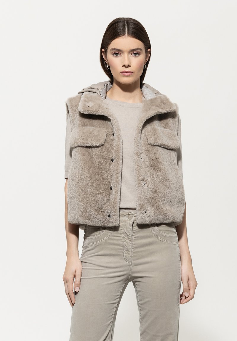 Frau mit glattem braunem Haar, die einen beigen Kurzarm-Pullover, eine taubengraue Kunstpelz-Weste und hellgraue, hochgeschnittene Hosen trägt.