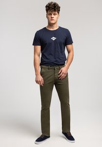 Granatowy bawełniany t-shirt z białym logo, zestawiony z oliwkowozielonymi chinosami oraz granatowymi tenisówkami z brązowymi sznurówkami, stojący na jasnym tle.