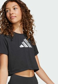 Crna skraćena majica s kratkim rukavima napravljena od glatkog materijala, s reflektirajućim, teksturiranim logotipom s tri pruge na prsima. Opusten kroj.