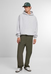 Šedá mikina s predným vreckom, vo voľnom strihu; olivovo zelené nohavice s viditeľnými švami; tmavozelená čiapka s bielym nápisom; biele tenisky.