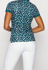 Polokošile s krátkým rukávem v tmavě zelenomodré barvě s vzorem bílých a černých tvarů. Látka je hladká, se stranovými rozparky na lemu.