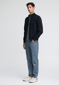 Svart zip-up jacka i stickad tyg, kombinerad med en grå skjorta under och ljusblå denimjeans. Skorna är beige med svarta accenter.